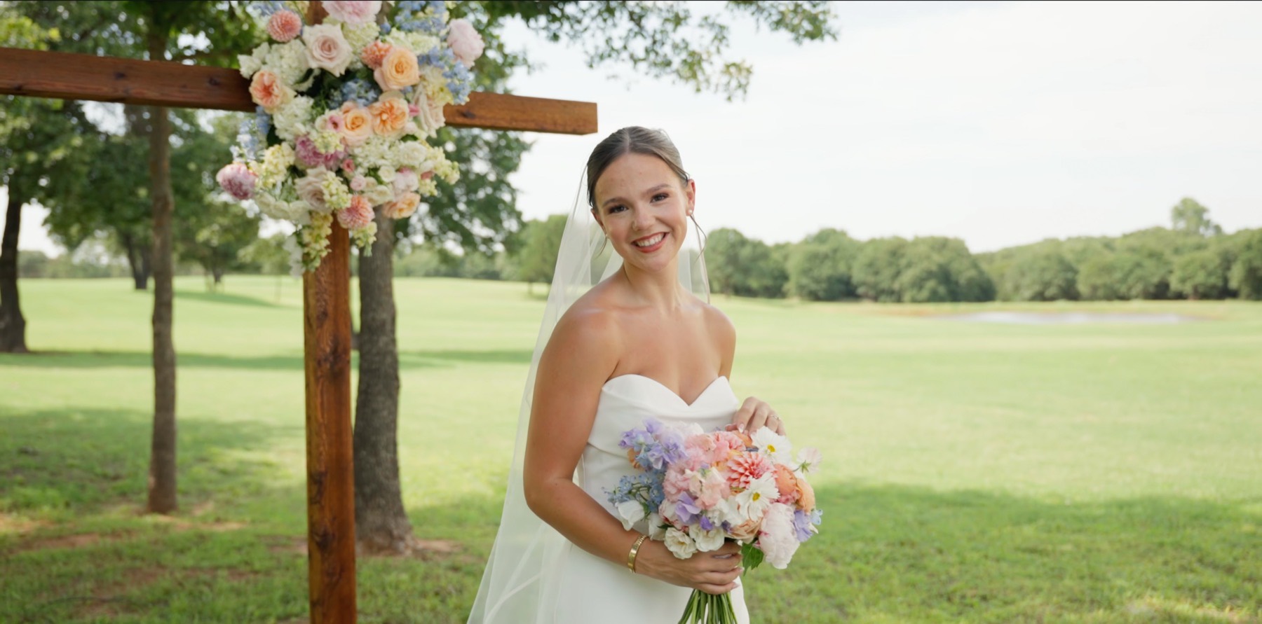 Bride smiling beneath a wooden arch of peach and blue flowers, an open green behind her