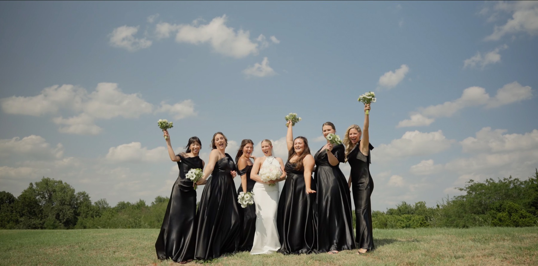 The bride with her bridesmaids in black, bouquets raised against a blue sky