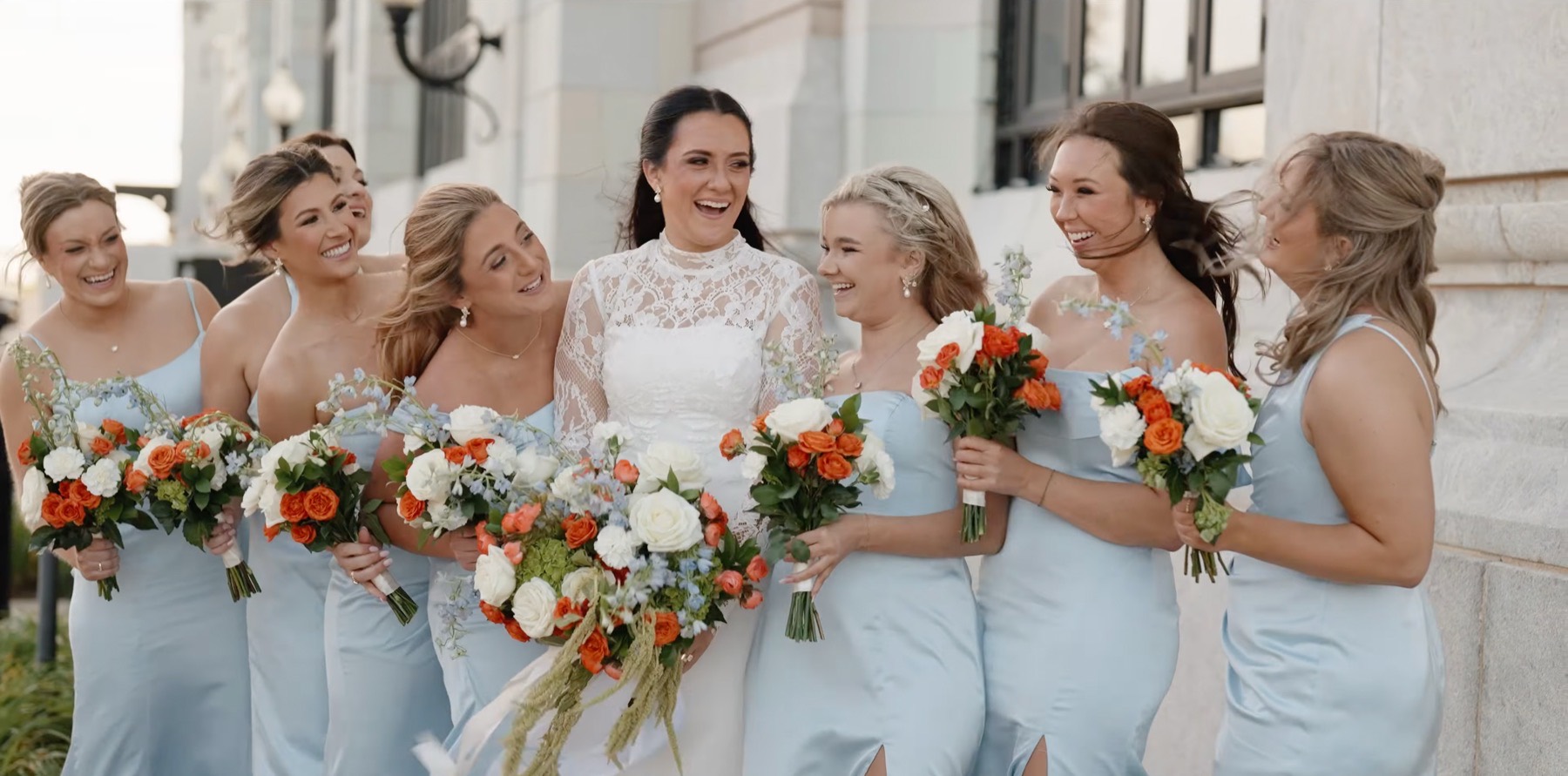 The bride laughing with her bridesmaids in pale blue, orange and white bouquets in their arms