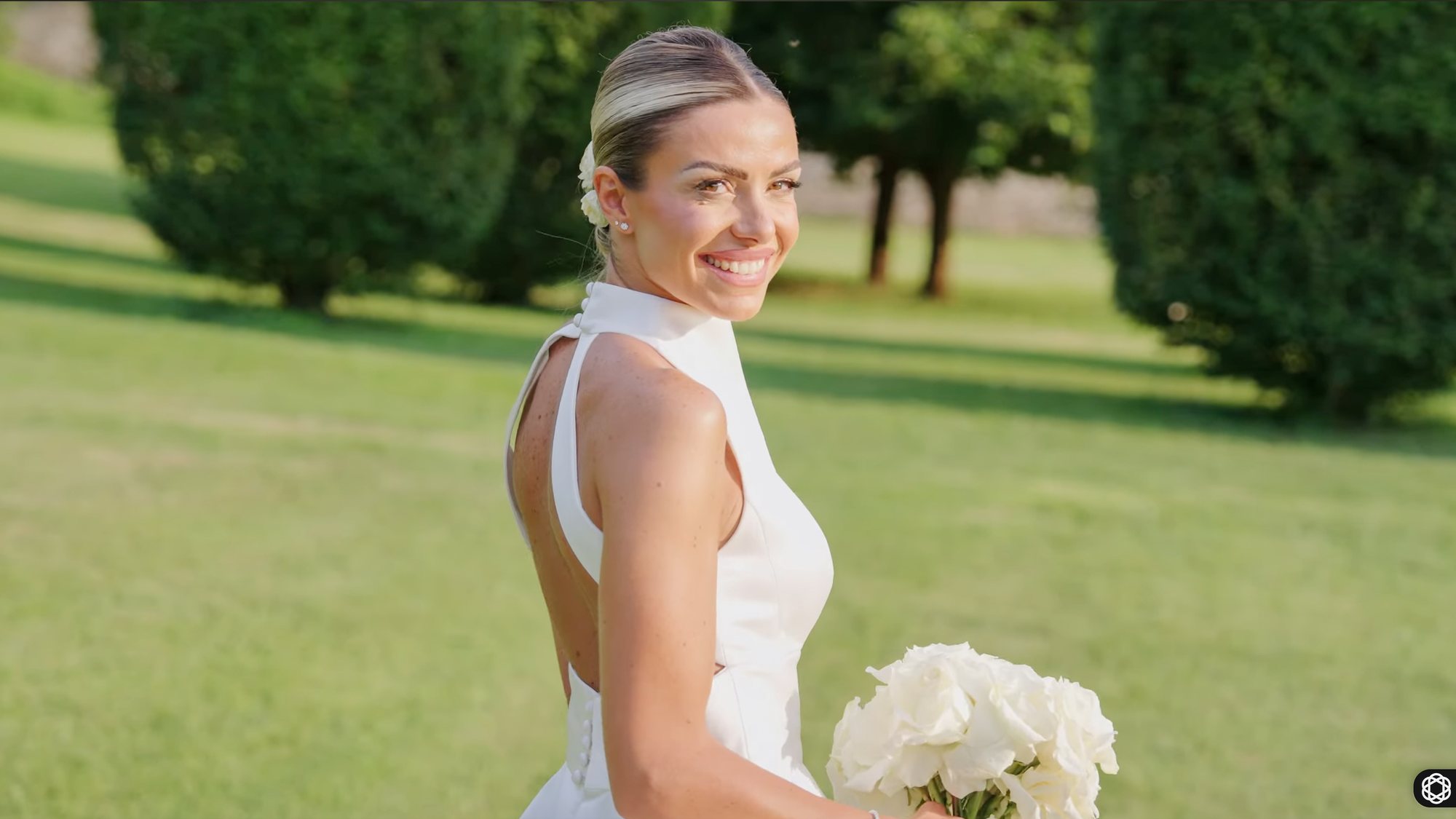 Bride in a white halter-neck gown holding a bouquet of white roses, looking back across a sunlit lawn