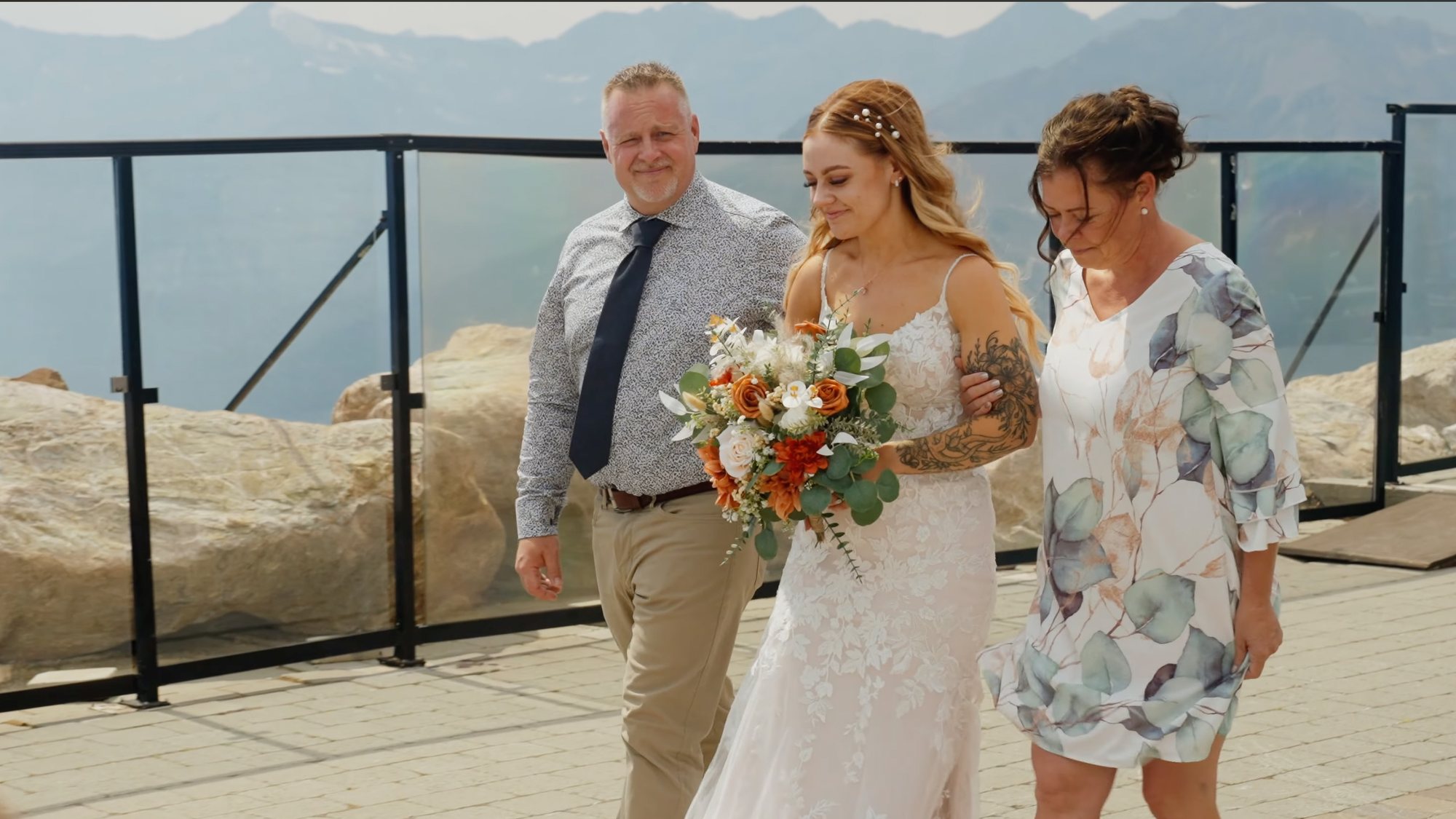 Bride walking between her parents along a mountain terrace, carrying an autumn bouquet