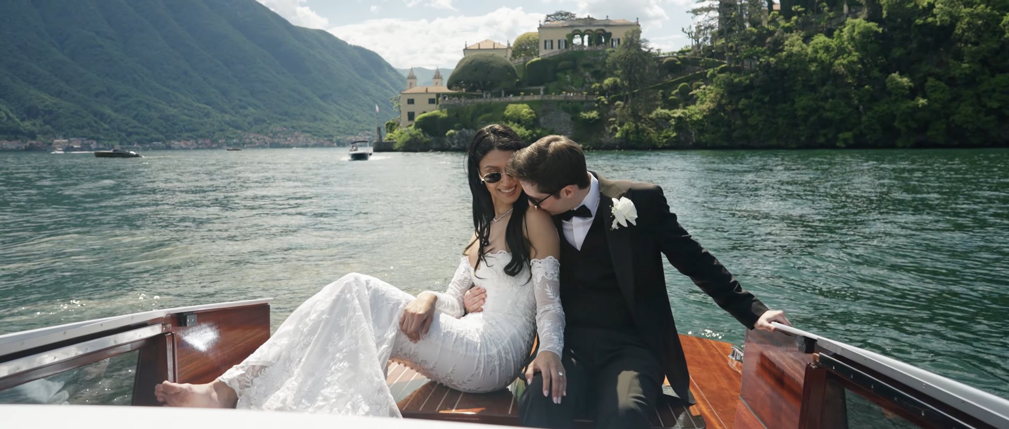 Bride and groom aboard a wooden boat on the lake, a villa on the far shore behind them