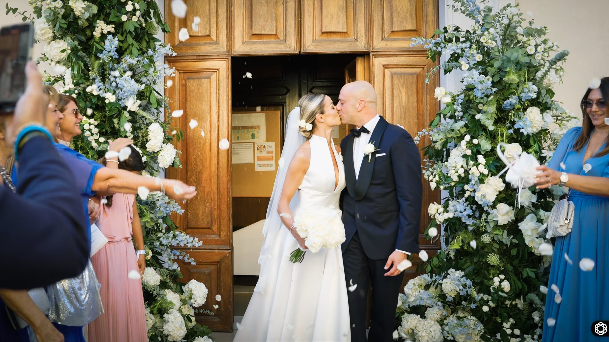 Bride and groom kissing in a church doorway beneath a floral arch as guests throw confetti