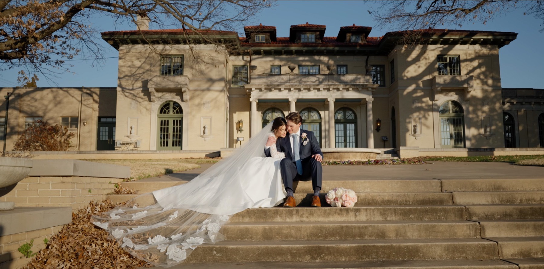 Bride and groom sitting on the steps of a stone mansion, her embroidered veil spread down the stairs