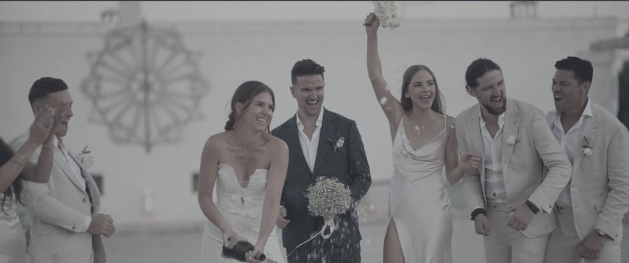 A wedding party laughing as the champagne is opened — the shot as the camera recorded it, flat and grey.